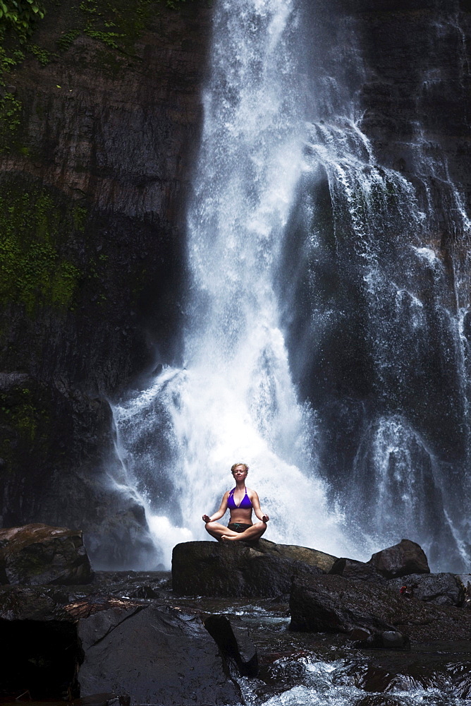 A beautiful young woman sitting in a lush jungle next to a waterfall in Lovina, Bali, Indonesia.