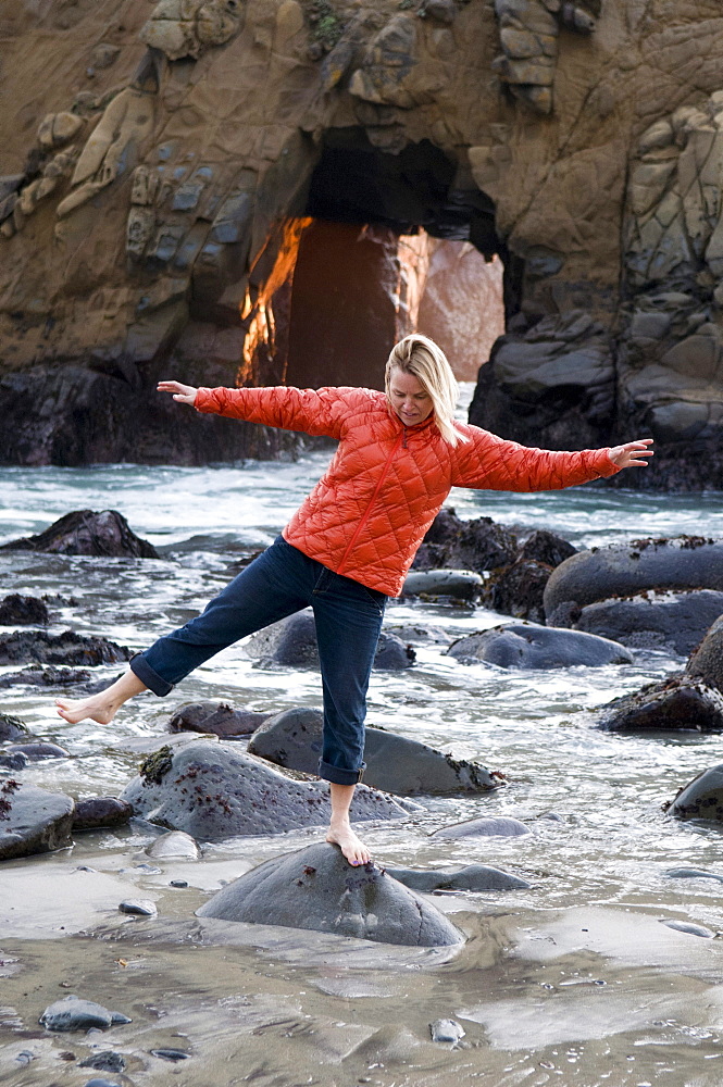 A woman balancing on a rockin front of a blow hole with the setting sun coming through, Pfeiffer Beach, Big Sur, California.