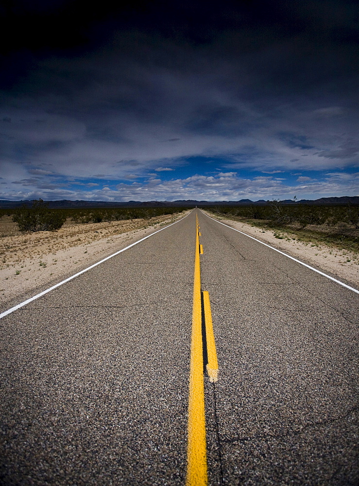 A road continues straight into the distance beneath a darkening sky in Mojave Desert, California.