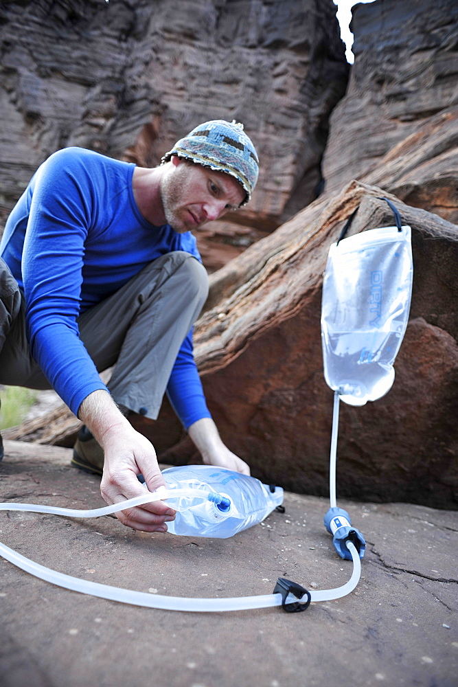 Male hiker filters water on a cliff-pinched patio near Deer Creek Falls in the Grand Canyon outside of Fredonia, Arizona November 2011.  The 21.4-mile loop starts at the Bill Hall trailhead on the North Rim and descends 2000-feet in 2.5-miles through Coconino Sandstone to the level Esplanada then descends further into the lower canyon through a break in the 400-foot-tall Redwall to access Surprise Valley.  Hikers connect Thunder River and Tapeats Creek to a route along the Colorado River and climb out Deer Creek.