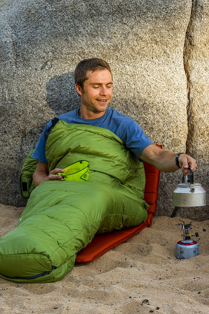 Man on the beach in a sleeping bag with a stove heating water.