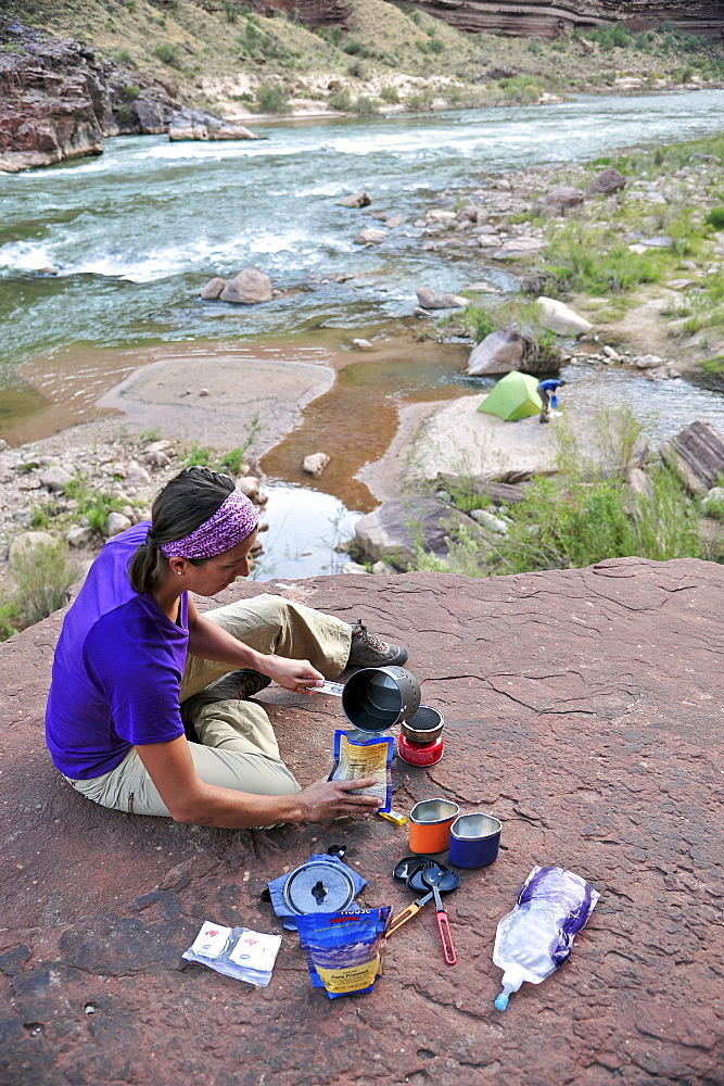 Female hiker cooks dinner on a cliff-pinched patio above camp and the Colorado River near Deer Creek Falls in the Grand Canyon outside of Fredonia, Arizona November 2011.  The 21.4-mile loop starts at the Bill Hall trailhead on the North Rim and descends 2000-feet in 2.5-miles through Coconino Sandstone to the level Esplanada then descends further into the lower canyon through a break in the 400-foot-tall Redwall to access Surprise Valley.  Hikers connect Thunder River and Tapeats Creek to a route along the Colorado River and climb out Deer Creek.