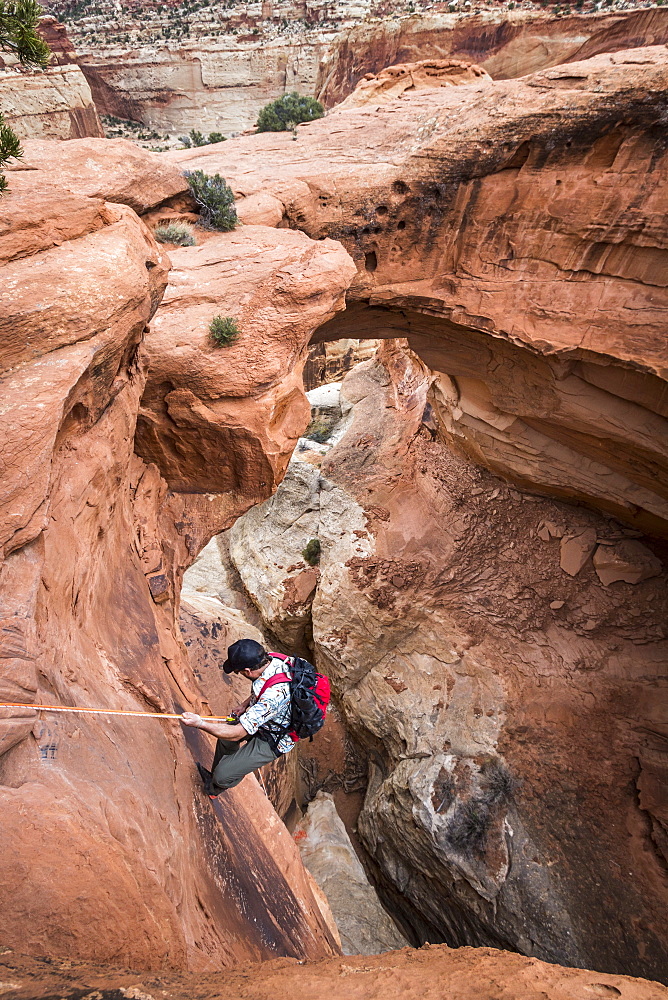 A man rappels down into a canyon below a natural rock arch in the desert.