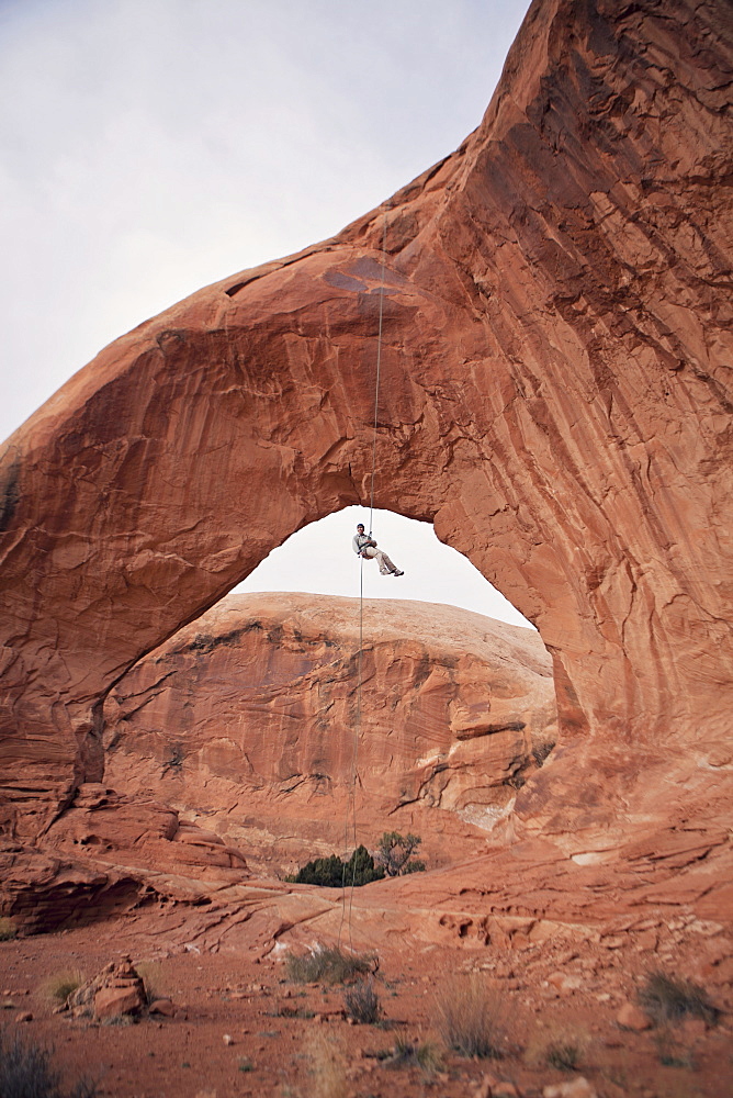 Rock climber hanging under eroded natural arch, Moab, Utah, USA