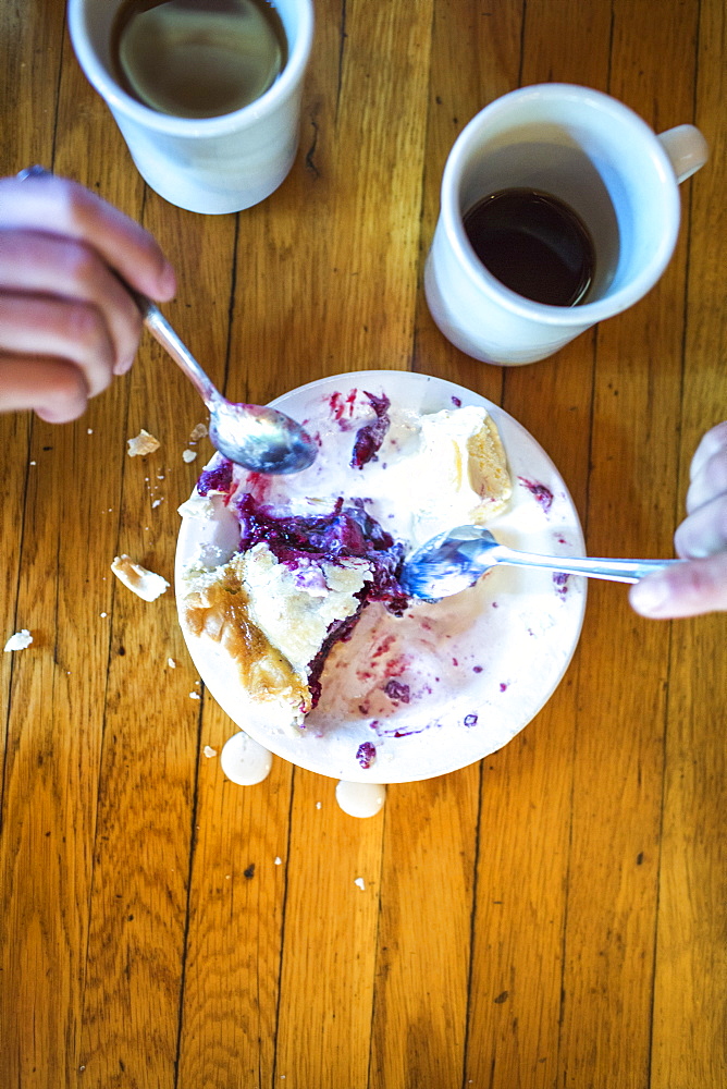 Two hands reach forks into a plate of pie and ice cream with two mugs of coffee next to the plate.