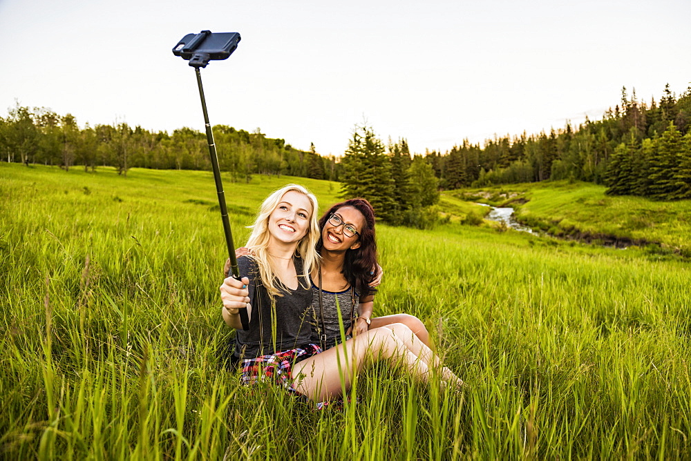 Two girlfriends sitting in a grass field posing for a self-portrait with a selfie stick and smart phone; Edmonton, Alberta, Canada