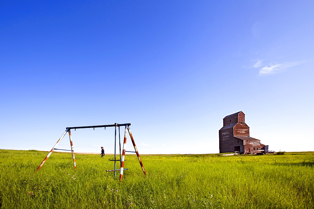 A Man Walking Through A Field With An Old Swing Set And An Old Grain Elevator, Bents, Saskatchewan, Canada