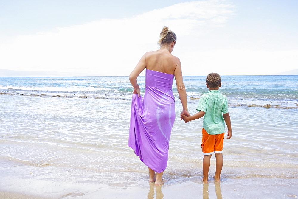 A mother and son hold hands while standing in the shallow water at the ocean, Hawaii united states of america