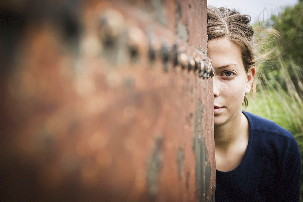 A young woman poses while peering out from behind a rusty weathered drum, South naknek bristol bay alaska united states of america