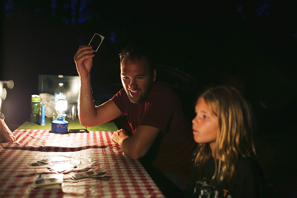 A Father And Daughter Playing Cards On A Picnic Table At Night By The Light Of A Lantern While Camping, Peachland, British Columbia, Canada