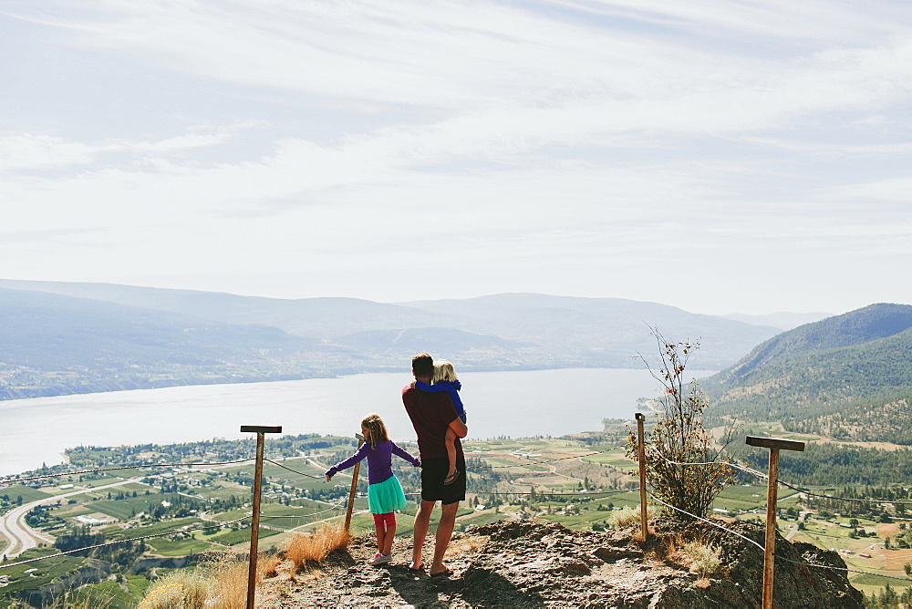 A Father With His Daughters Standing On A Rock Ledge Overlooking Lake Okanagan, Peachland, British Columbia, Canada