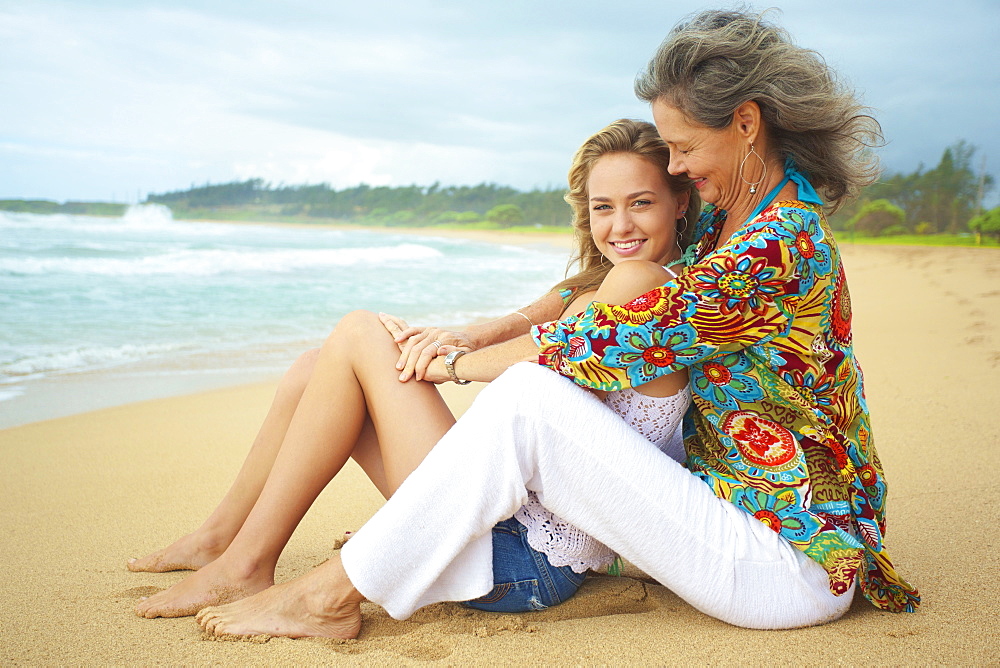 A Mother And Daughter Sitting Together On The Beach At The Water's Edge, Kauai, Hawaii, United States Of America