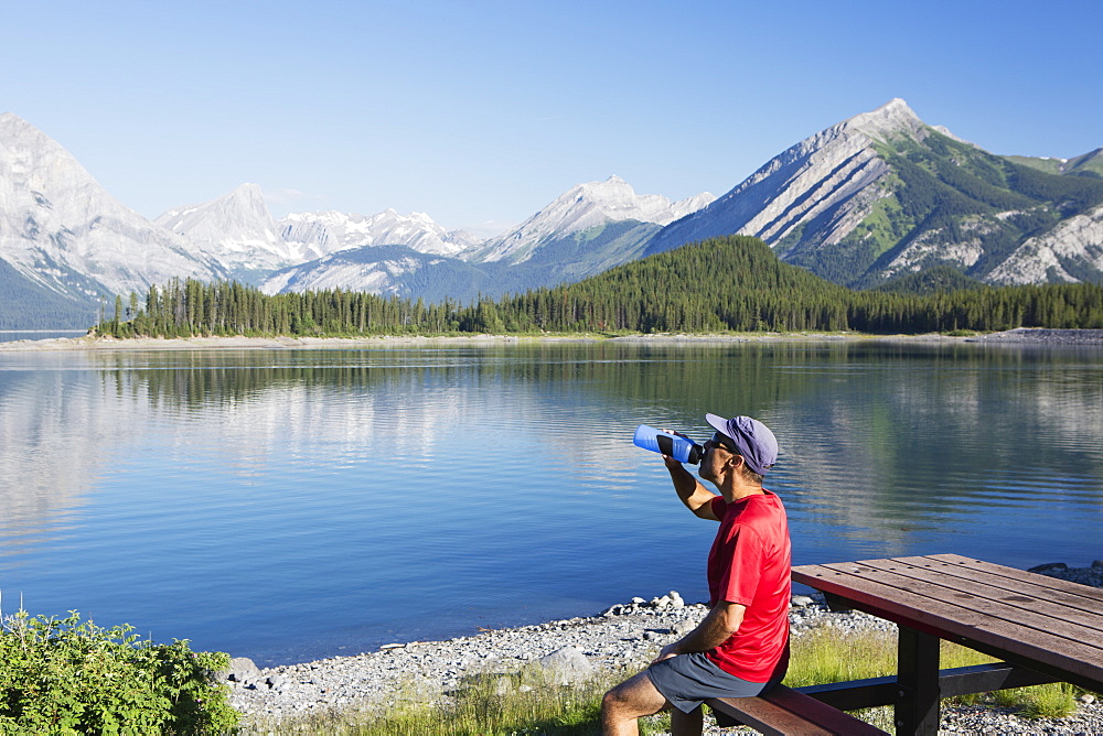 A Man Drinks From A Water Bottle On The Edge Of A Lake Overlooking The Rocky Mountains, Kananaskis, Alberta, Canada