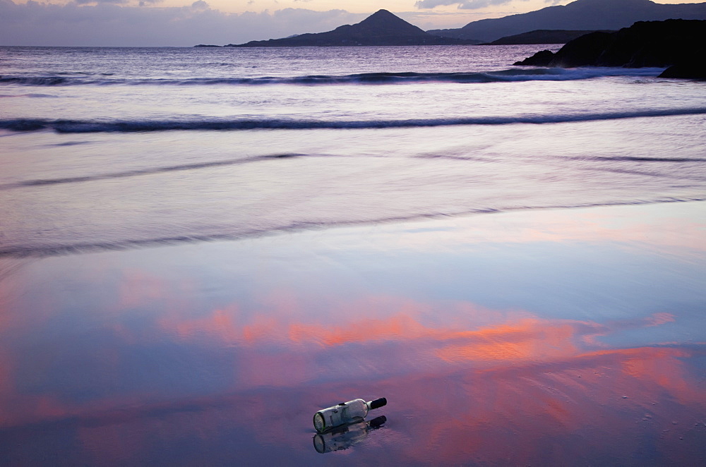 A Glass Bottle Lays On The Wet Sand At The Water's Edge At Sunset, County Kerry, Ireland