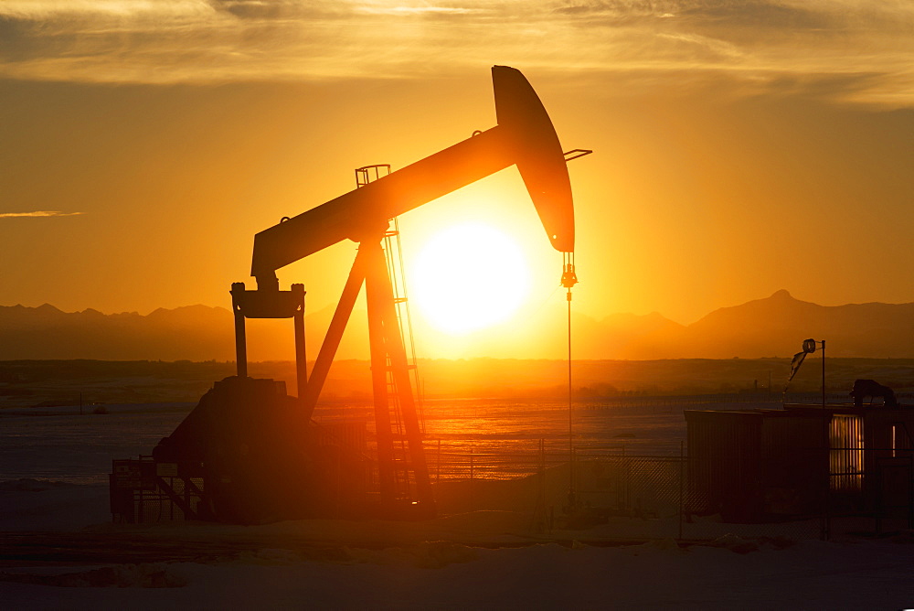 Silhouetted Pumpjack At Sunset With Mountains In The Background And Glowing Sunlight, Alberta, Canada