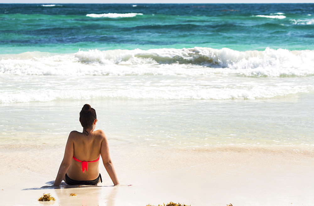 Young Woman Sitting On Sandy Beach In Water Looking Out At Waves Coming In And Blue Sky, Akumal, Quintana Roo, Mexico