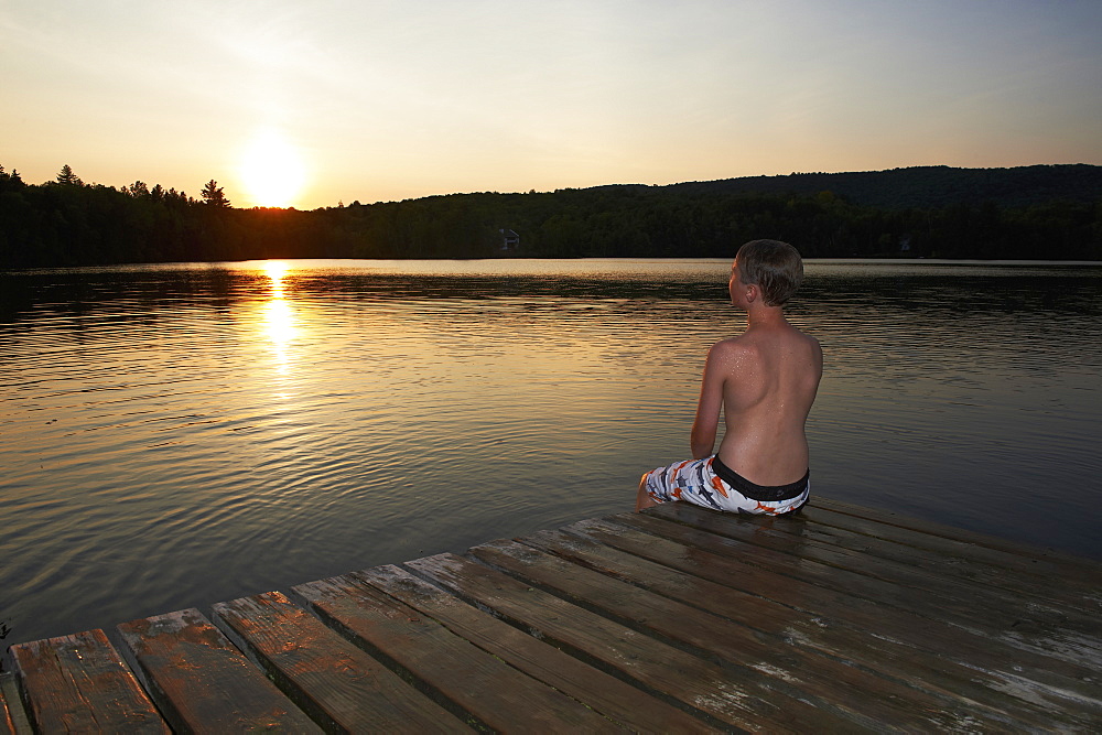 Boy Sitting On The Dock With Feet In Water At Sunset, Lac Des Neiges, Quebec, Canada