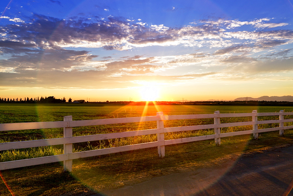 Sunlight Shining Across A Grass Field And White Rail Fence At Sunset, British Columbia, Canada