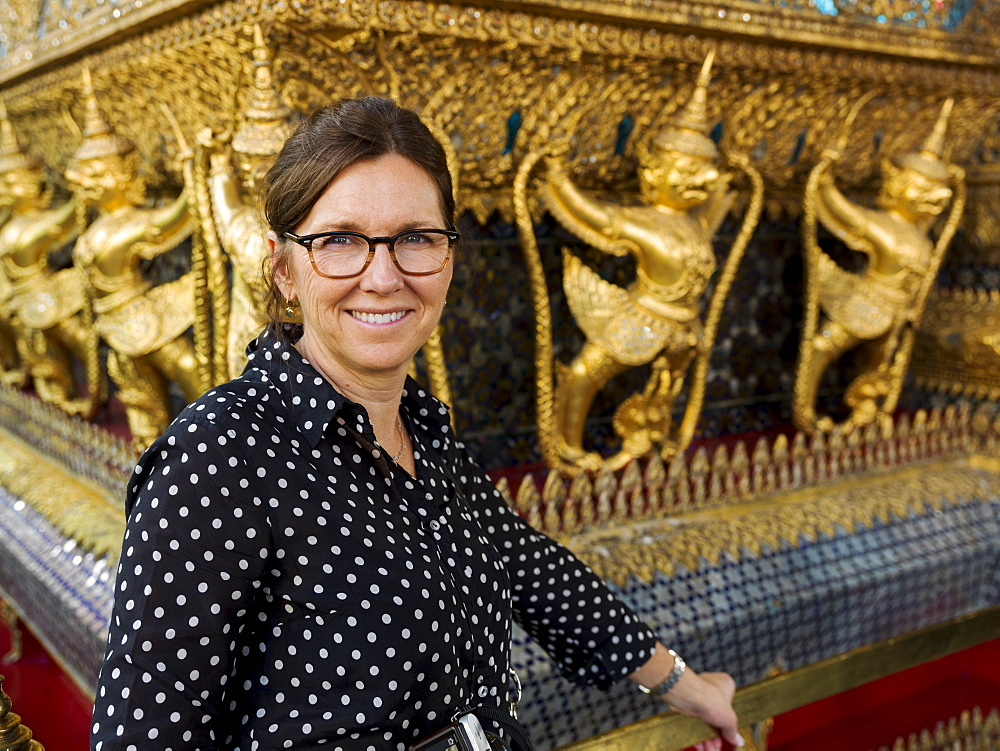 Portrait Of A Woman Standing Beside A Wall With Gold Statues, Temple Of The Emerald Buddha (Wat Phra Kaew), Bangkok, Thailand