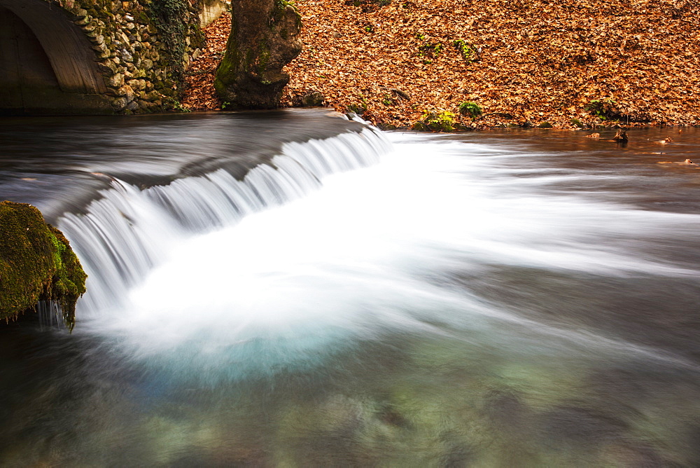 Water Cascading Over A Ledge In A River With The Riverbank Covered With Autumn Coloured Fallen Leaves, Naoussa, Greece