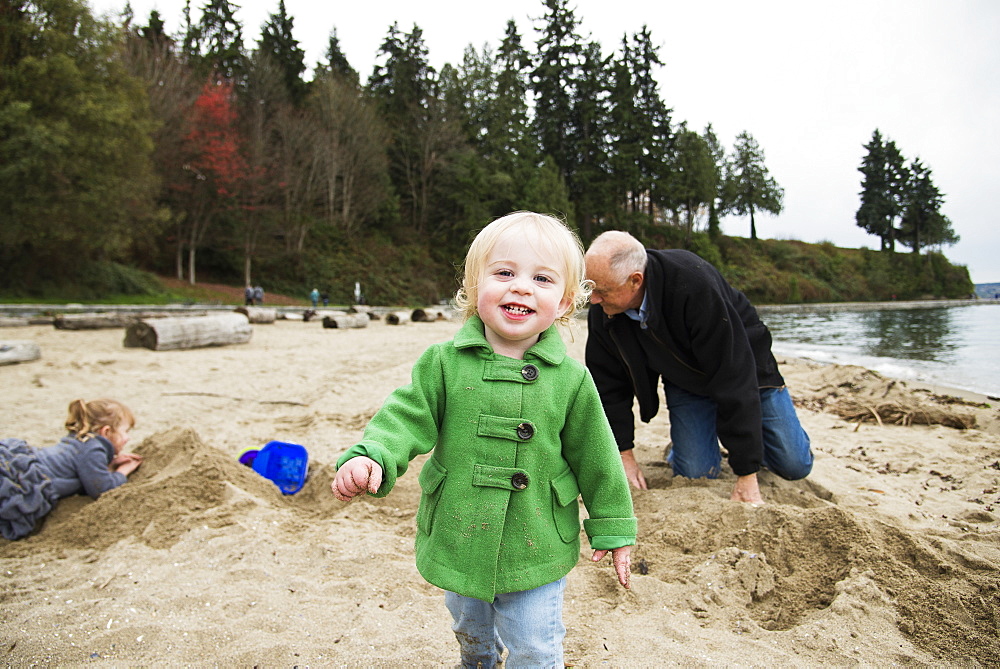 A Young Girl Walks Towards The Camera While A Grandfather And Granddaughter Play In The Background On The Beach In Winter At Stanley Park, Vancouver, British Columbia, Canada