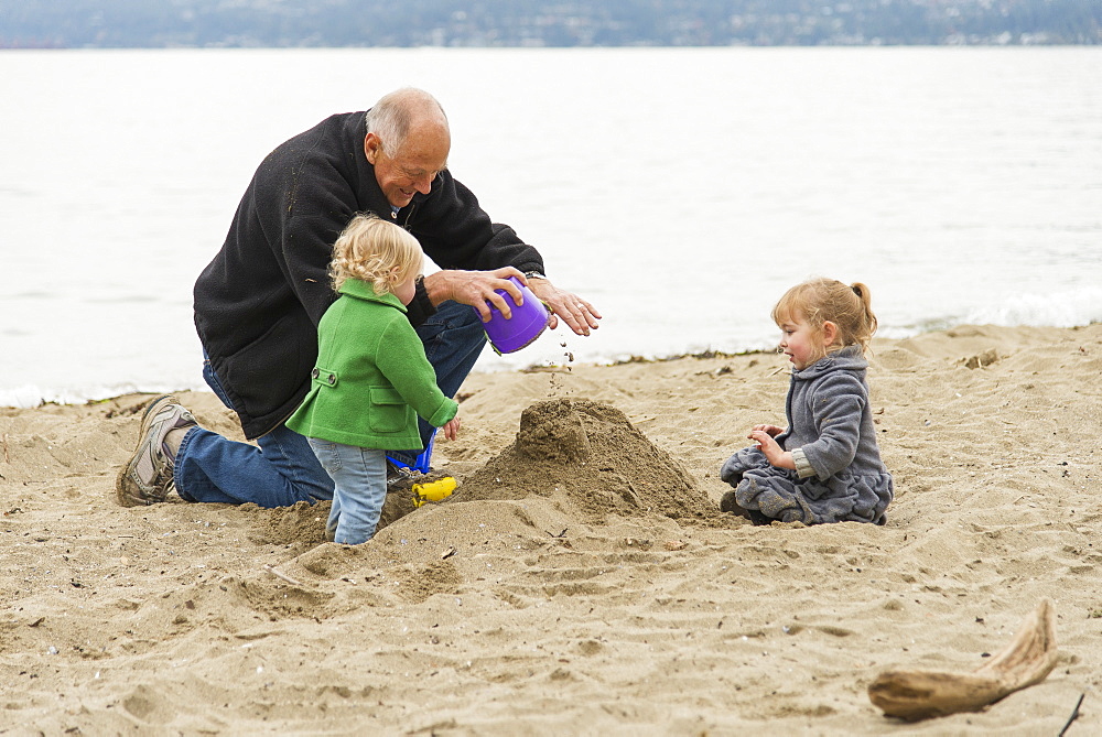 In Cold Weather, A Grandfather Plays With His Two Grand-Daughters On The Beach At Stanley Park, Vancouver, British Columbia, Canada