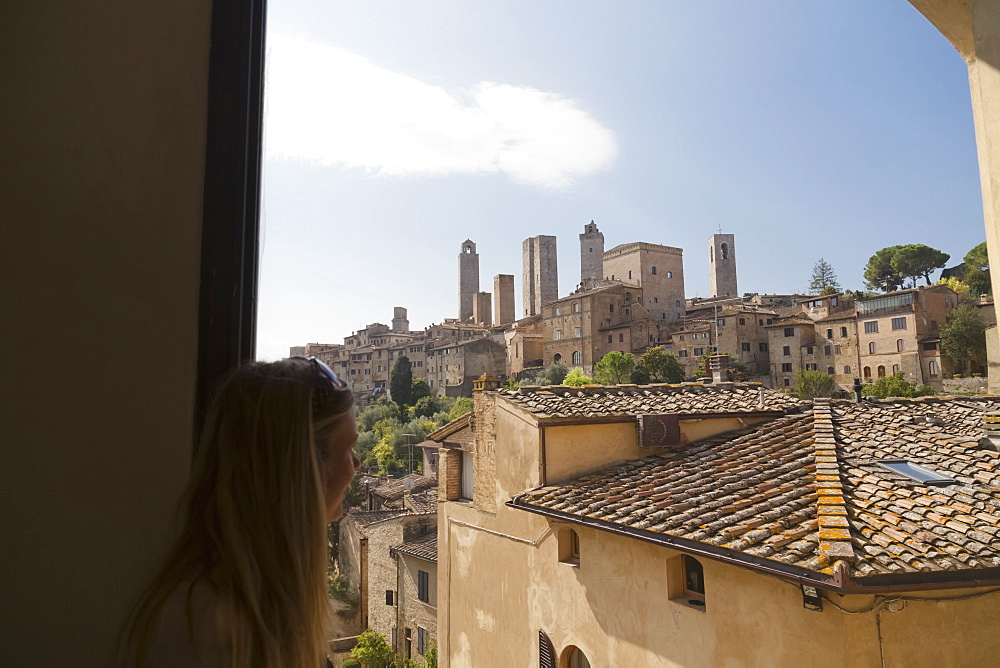 A Female Tourist Looks Out The Window At The Walled Medieval Village Of San Gimignano, San Gimignano, Province Of Siena, Tuscany, Italy