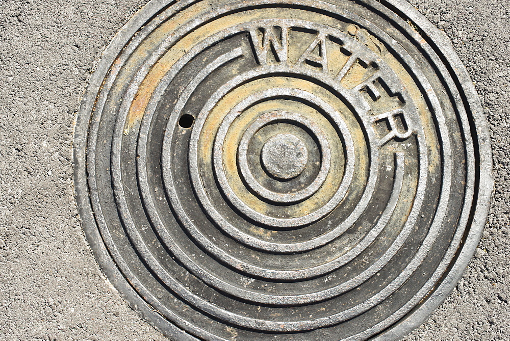 The Hot Sun Beats Down On A Manhole Cover With The Word 'water' Engraved On It, Waikaloa, Island Of Hawaii, Hawaii, United States Of America