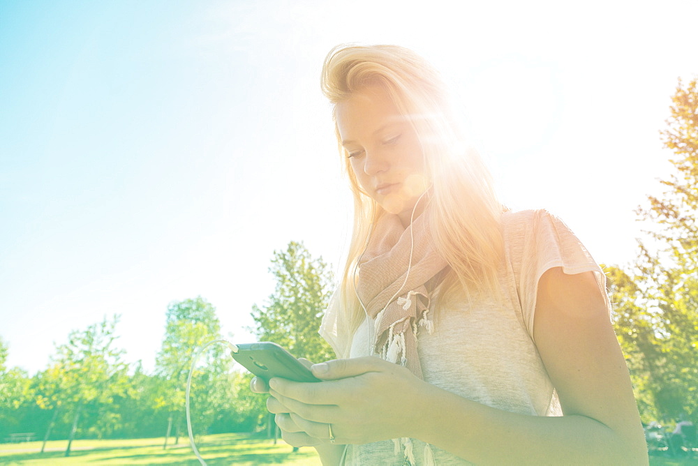 Young Woman With Long Hair Listening To Music Outdoors On Her Smart Phone In A City Park In Autumn, Edmonton, Alberta, Canada