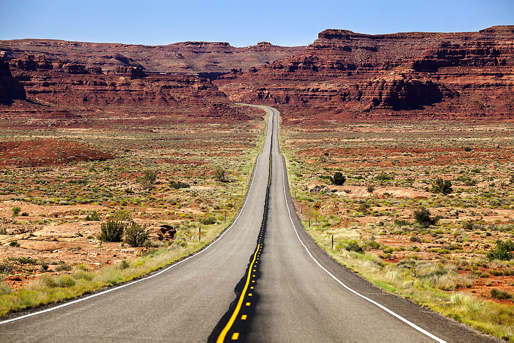 Utah Highway Stretching Into The Distance With Rock Cliffs And Blue Skies In The Background And Desert Vegetation On Either Side Of The Road, Utah, United States Of America