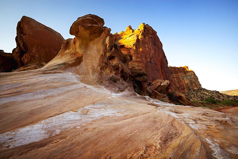 Smooth, Worn Rock Formations In The Foreground Sweeping Up To Tall Canyon Cliffs In The Background Under Clear Blue Skies, Capitol Reef National Park, Utah, United States Of America