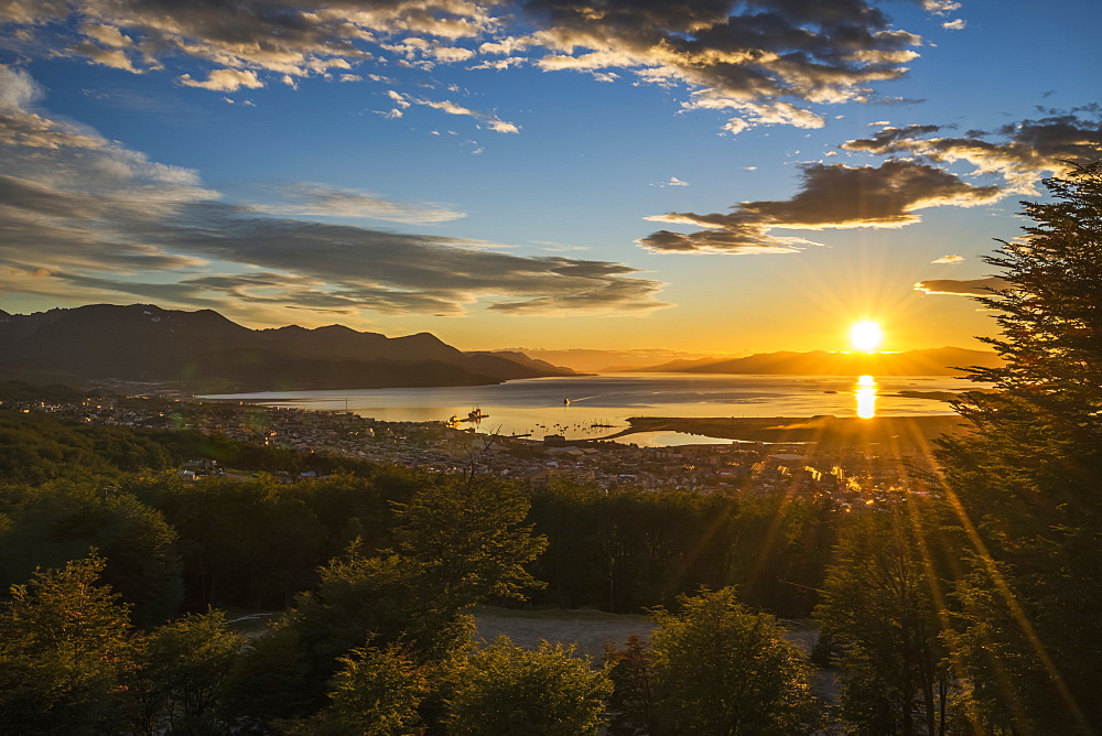 The Sun Rises Over The Port Of Ushuaia In Argentina As A Ship Enters The Calm Bay, Trees In The Foreground And Low Hills On The Horizon, While The Sky Is Dotted With Clouds And Turns From A Golden Yellow To A Deep Blue, Ushuaia, Argentina