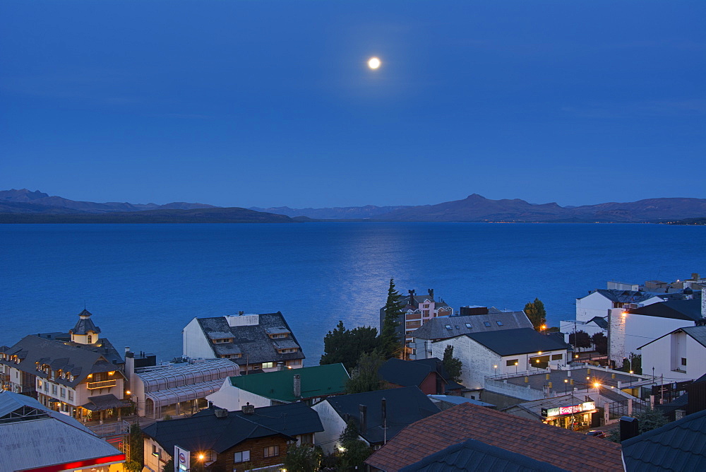 The Moon Rising Over A Lake From Above The Town Roof Tops Showing Some Yellow City Lights Against The Blue Scene, Bariloche, Argentina