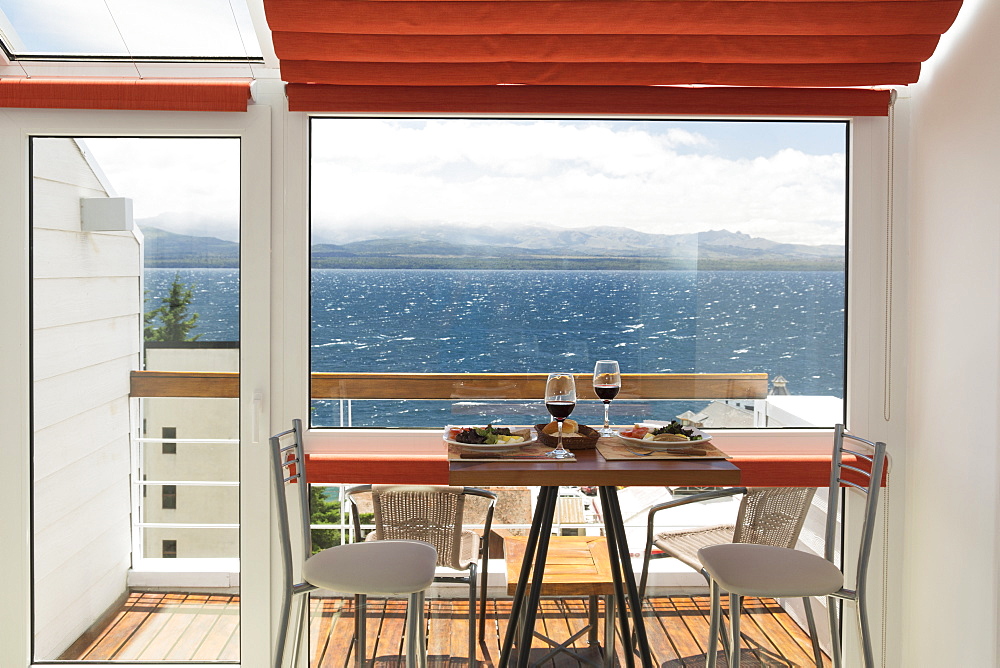 A Lunch Table With Two Glasses Of Red Wine Is Set In Front Of The Balcony Window Of A Bright Apartment With View Over A Lake, Bariloche, Argentina