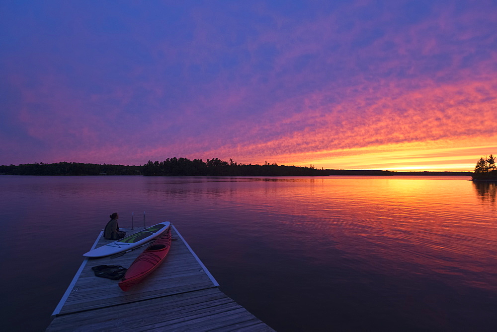 A Young Woman Sits At The End Of The Dock With Kayaks Watching The Sun Set Over A Lake, Ontario, Canada