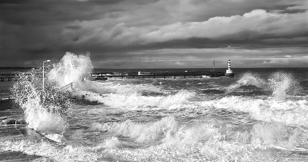 Waves Crashing Along The Coast With A Small Lighthouse At The End Of A Pier, Amble, Northumberland, England