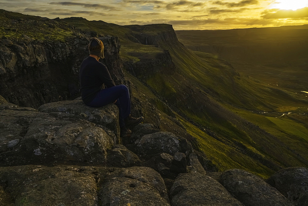 Person Sitting On A Cliff Top Overlooking The Sunset Near The Town Of Djupavik, Iceland