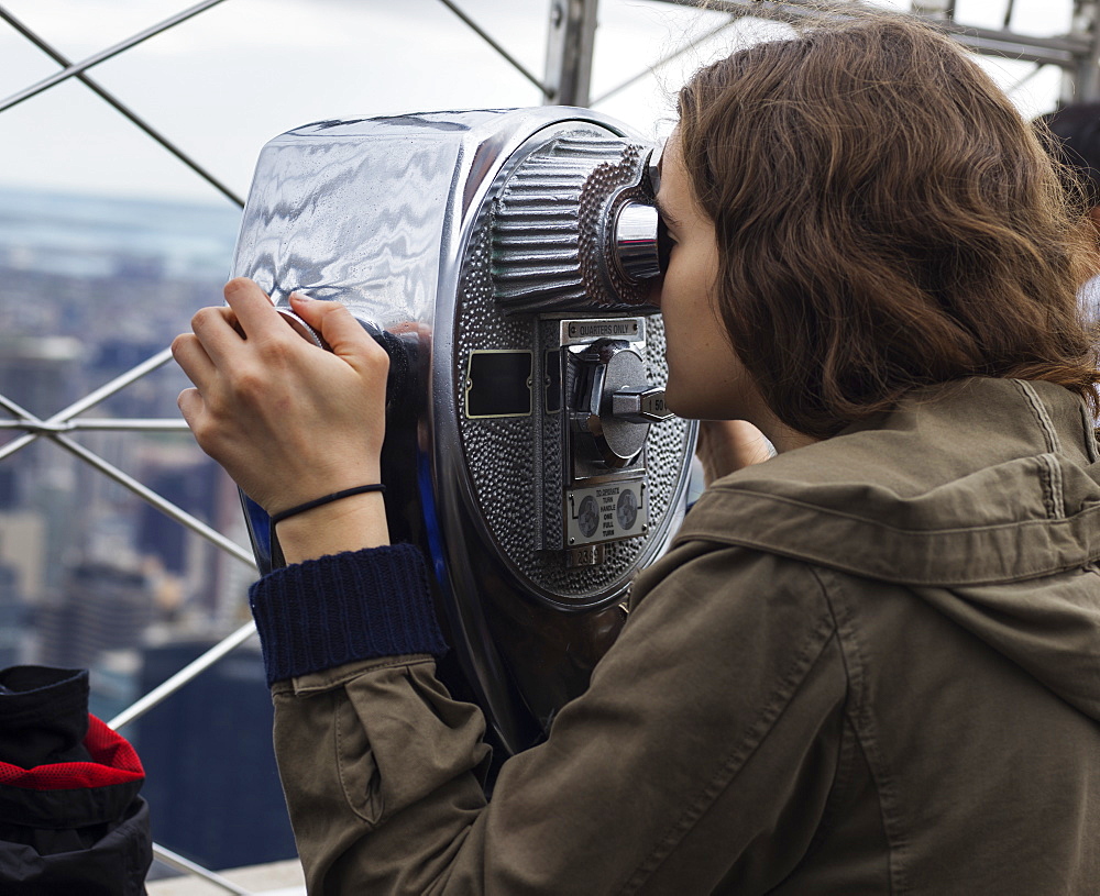 A Young Woman Looks Through Binoculars On The Observation Deck Of The Empire State Building To A View Of The Cityscape Of New York City, New York City, New York, United States Of America