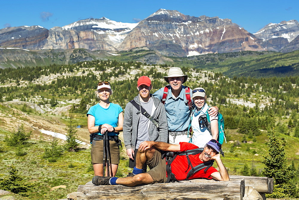 Group Of Hikers Posing On A Log Bench With Valley Below And Mountains And Blue Sky In The Distance, Banff, Alberta, Canada