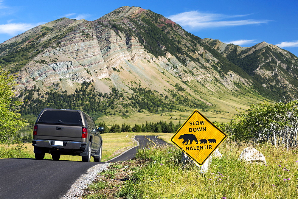 Cautionary Road Sign Of Bear Crossing On A Windy Mountain Road With Vehicle On Roadway, With Mountains And Blue Sky, Waterton, Alberta, Canada