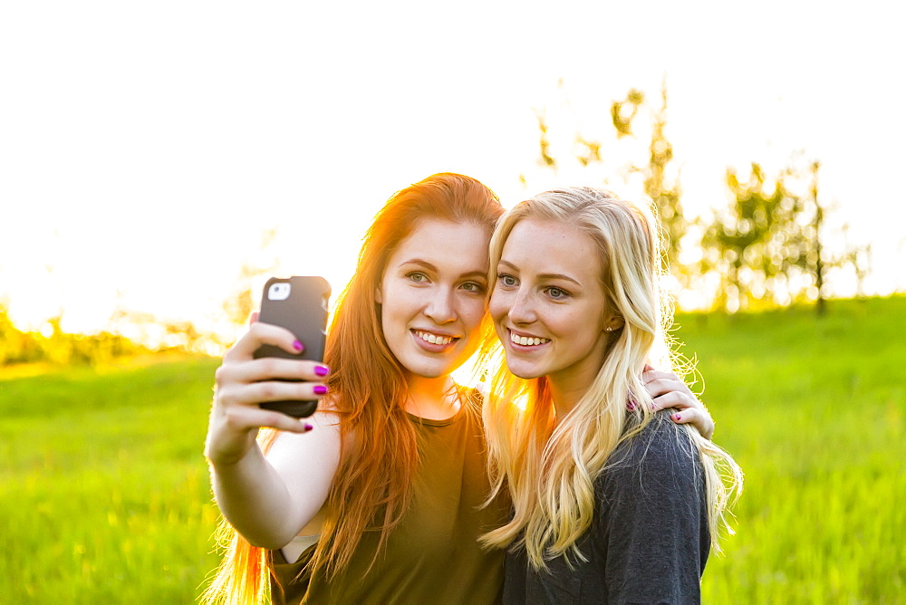 Two friends standing in a park taking a self-portrait with a smart phone at dusk, Edmonton, Alberta, Canada