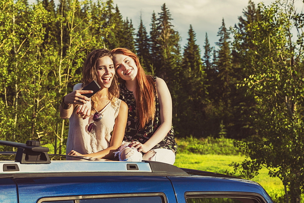 Two young women on a road trip stand up in the sunroof of a vehicle taking a self-portrait with a smart phone, Edmonton, Alberta, Canada