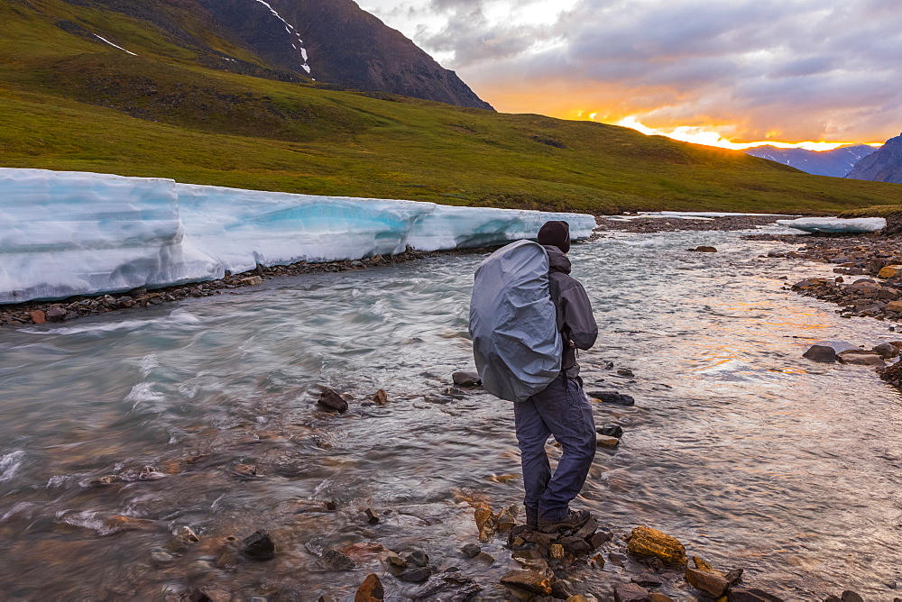 A backpacker observes the midnight sun peeking through the clouds from an unnamed fork of the Atigun River still partially covered in aufeis (sheet-like ice formations) in a remote valley of the Brooks Range, Alaska, United States of America