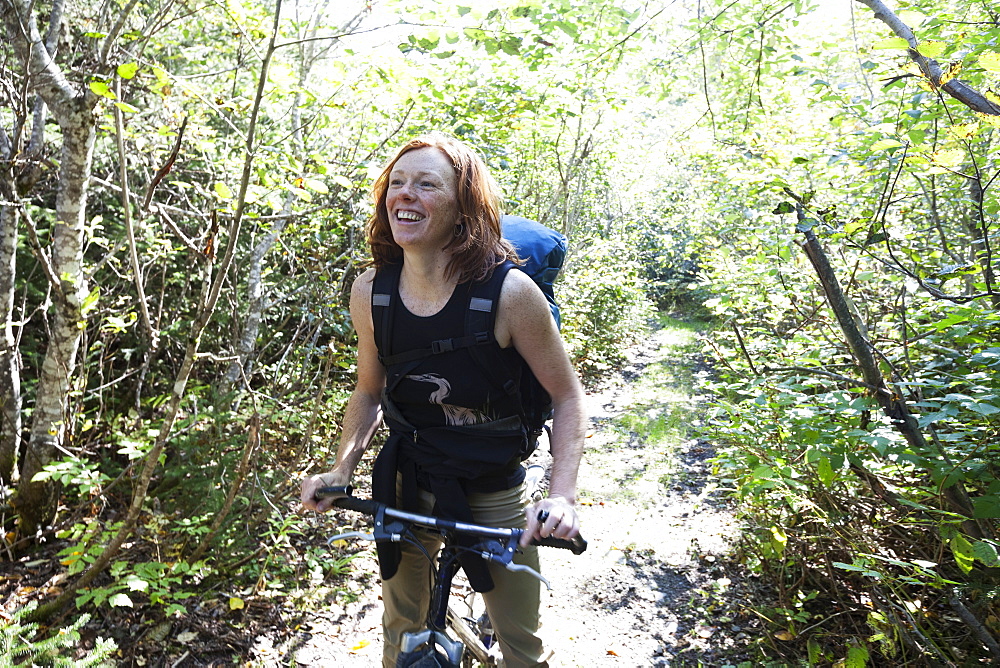 A Woman Stands On A Path In A Forest With Her Bicycle And A Backpack, Alaska, United States Of America