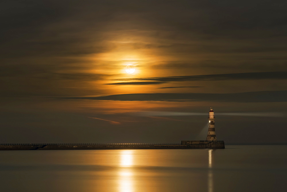 Roker Lighthouse Casts A Light With The Golden Sunlight Shining Through Cloud And Reflecting On Tranquil Water, Sunderland, Tyne And Wear, England