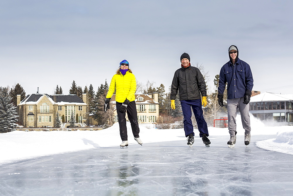 Two Males And One Female Skating On Freshly Groomed Ice On Pond With Houses In The Background, Calgary, Alberta, Canada