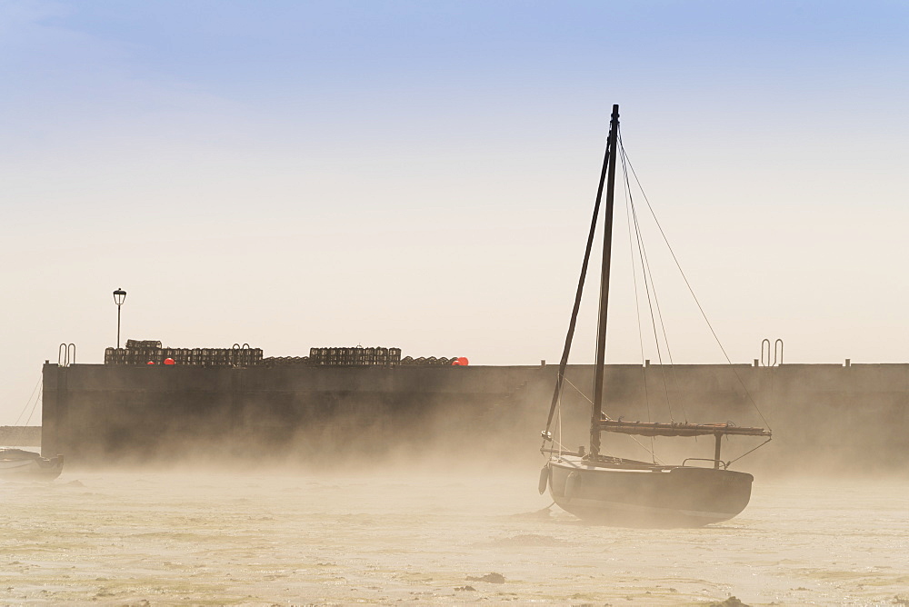 Wind Blows Sand Up On The Beach Against A Sailboat Sitting On The Shore, Lindisfarne, Northumberland, England