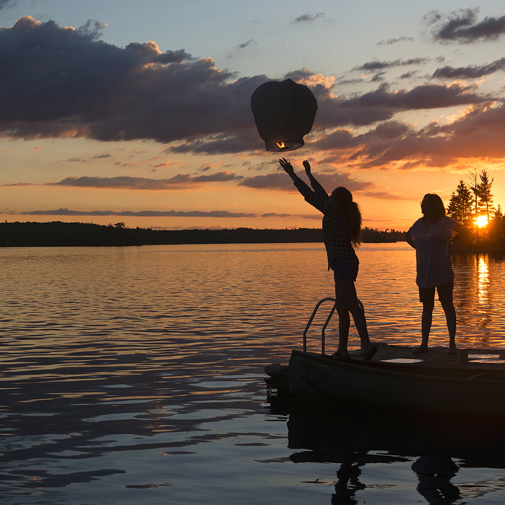 Silhouette Of A Teenage Girl Releasing A Lit Lantern Out Over A Lake At Sunset, Lake Of The Woods, Ontario, Canada