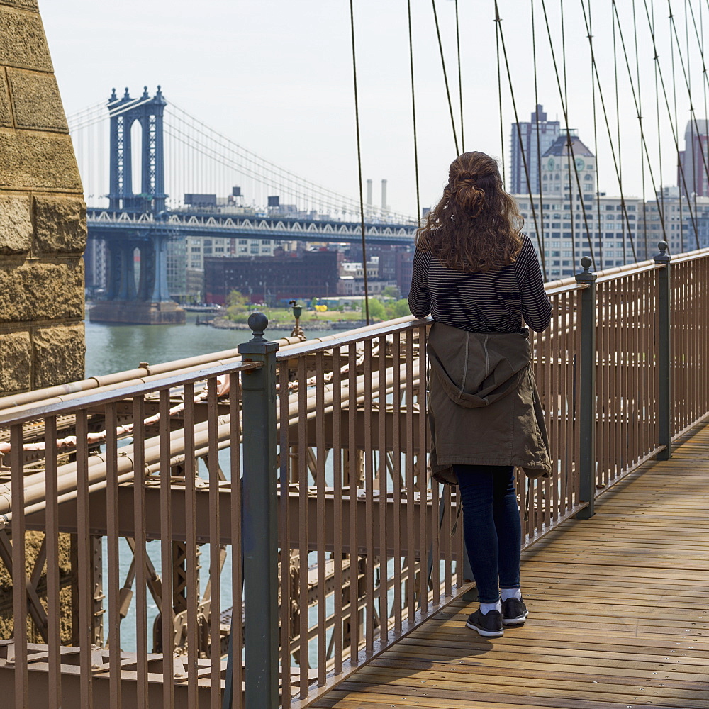 A Young Woman Stands On The Brooklyn Bridge Looking Out Over The East River To The Manhattan Bridge, New York City, New York, United States Of America