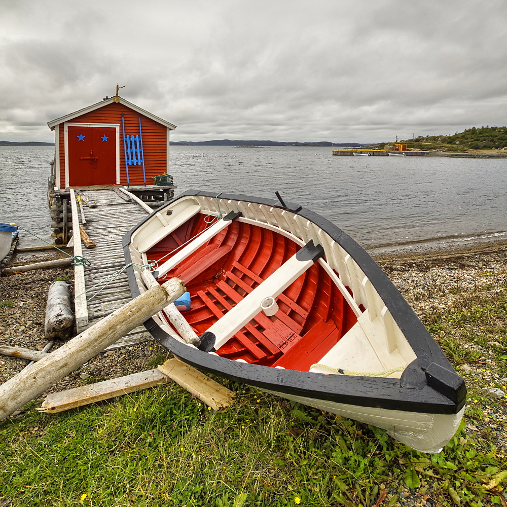 A red and white canoe lays on the shore with a red fishing shed along the water's edge of the Atlantic coast, Newfoundland, Canada